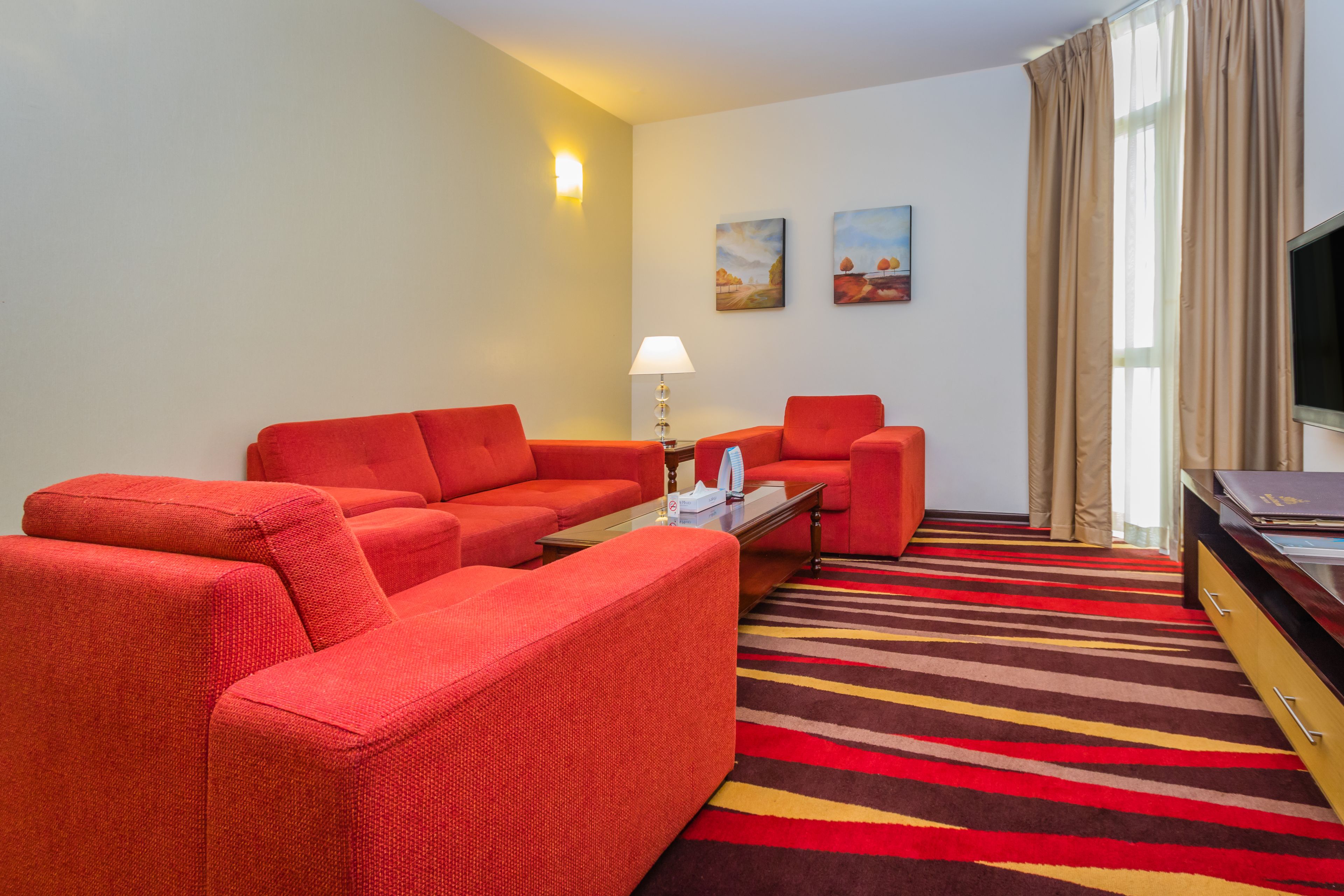 A hotel room with a red sofa set, a small table with a lamp, a TV, and a striped carpet.