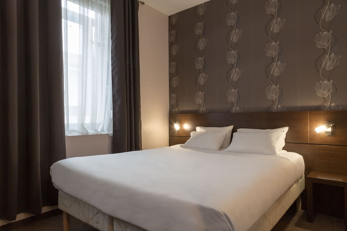 A neatly made bed with white linens, a headboard with wall-mounted lights, and a window with sheer curtains.