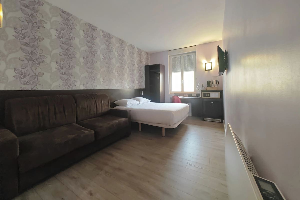 A small, furnished room with a sofa, a bed, a desk, and a window.