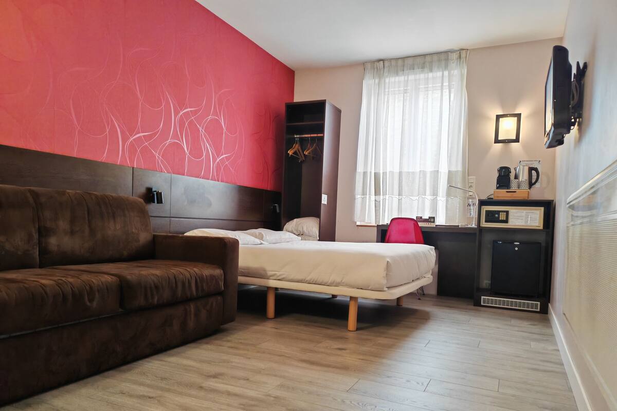 A room with a brown sofa, a bed with white bedding, a red accent wall, a window with sheer curtains, a small desk with a computer, and a wall-mounted TV.