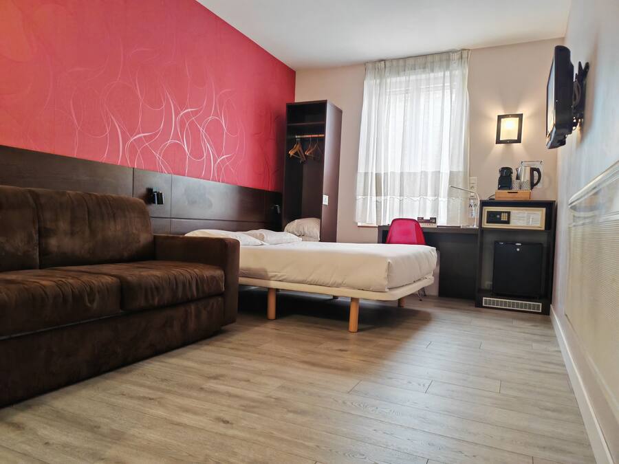 A room with a brown sofa, a bed with white bedding, a red accent wall, a window with sheer curtains, a small desk with a computer, and a wall-mounted TV.