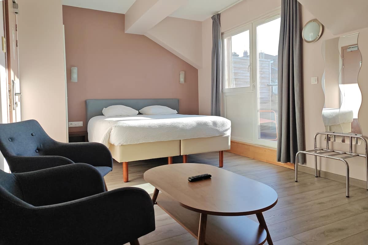 A modern hotel room with a bed, two armchairs, a small table, and a window with curtains.
