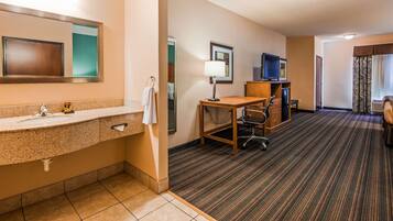 A hotel room with a bathroom, a desk, a chair, a television, and a carpeted floor.