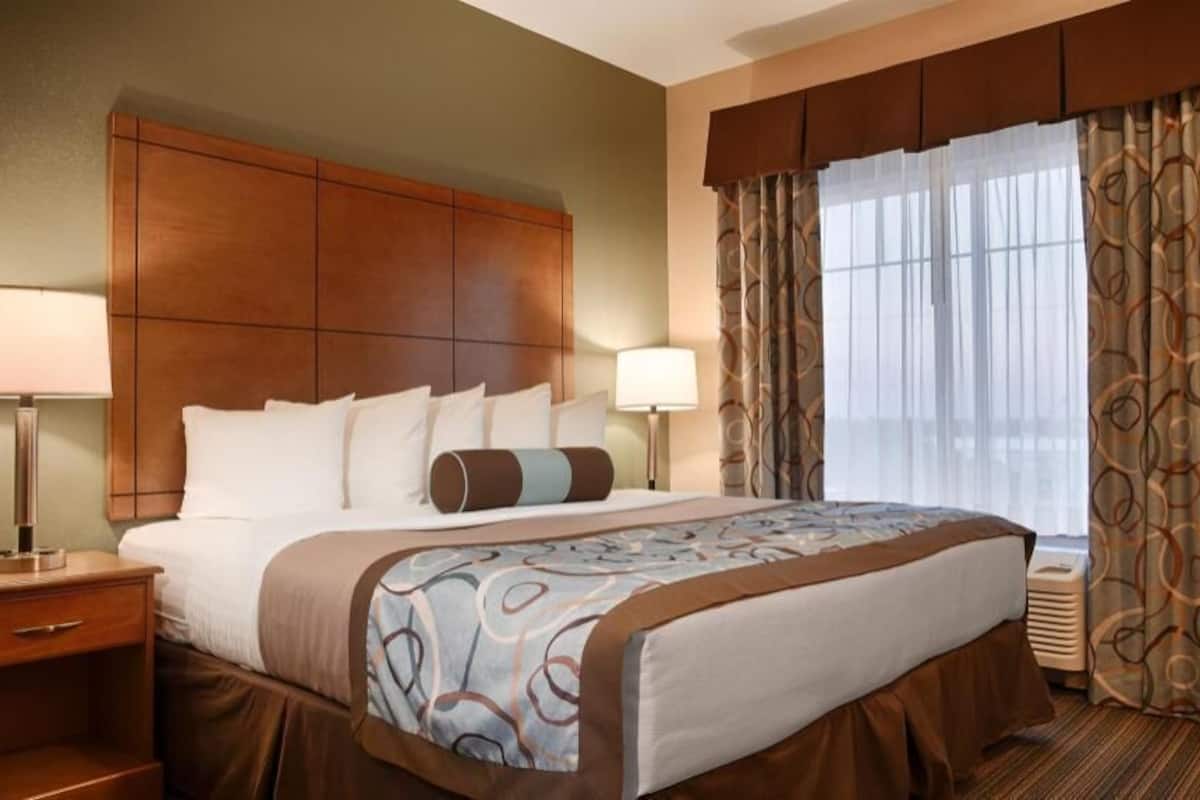 A hotel room with a large bed, two bedside tables with lamps, a headboard, a window with curtains, and a wall-mounted heater.