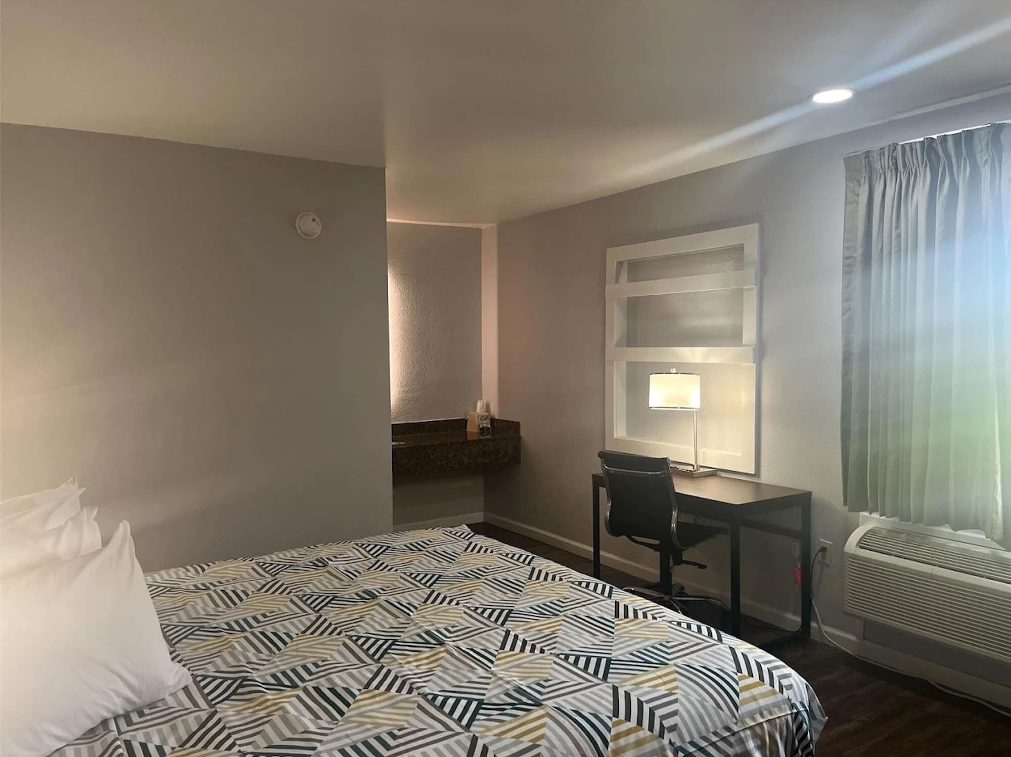 Ein Hotelzimmer mit Bett, Schreibtisch, Stuhl, einem Fenster mit Vorhängen und einer Wandklimaanlage.