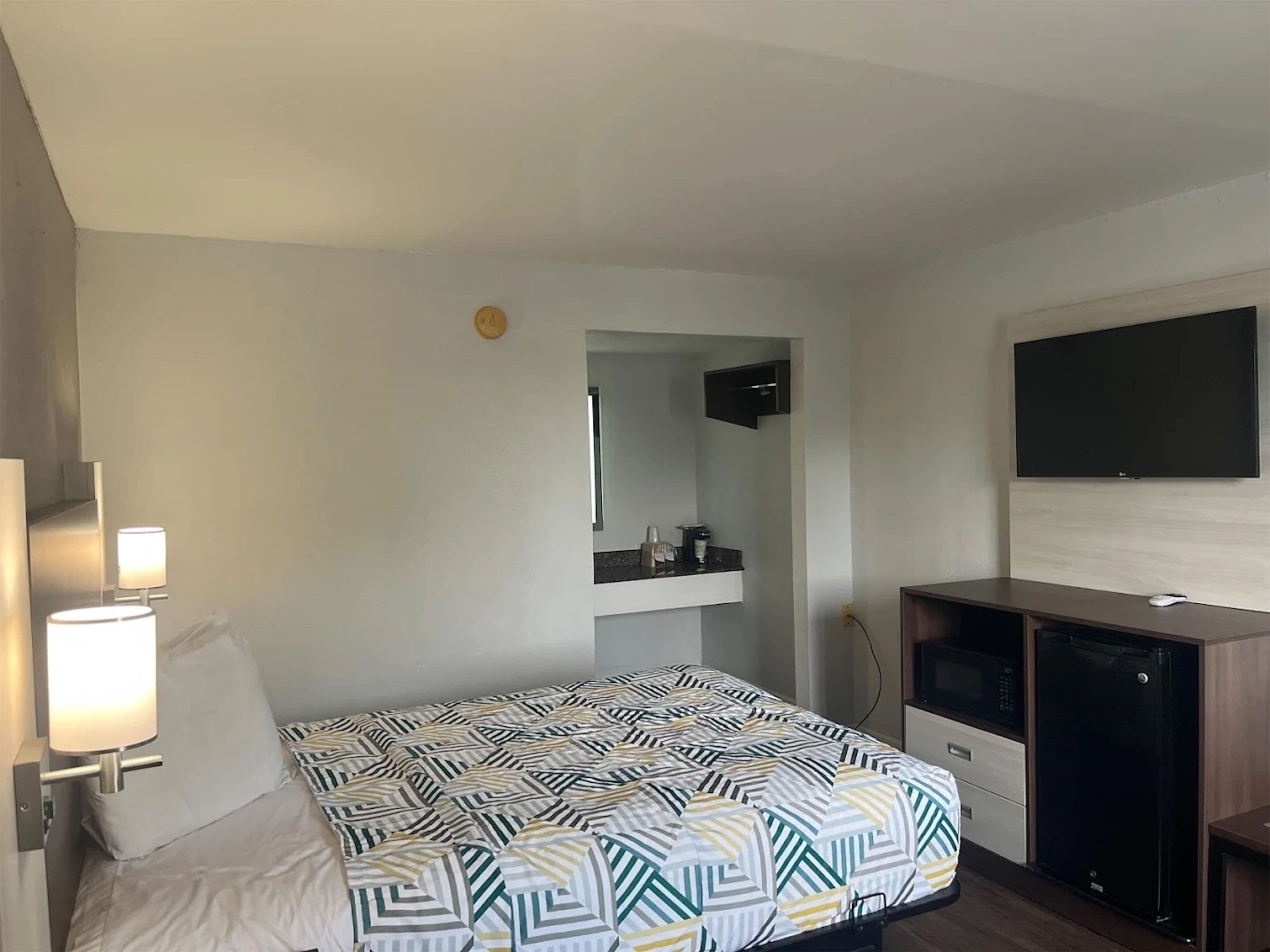 Habitación de hotel con cama, lámparas de noche, televisión, kitchenette pequeña y espejo.