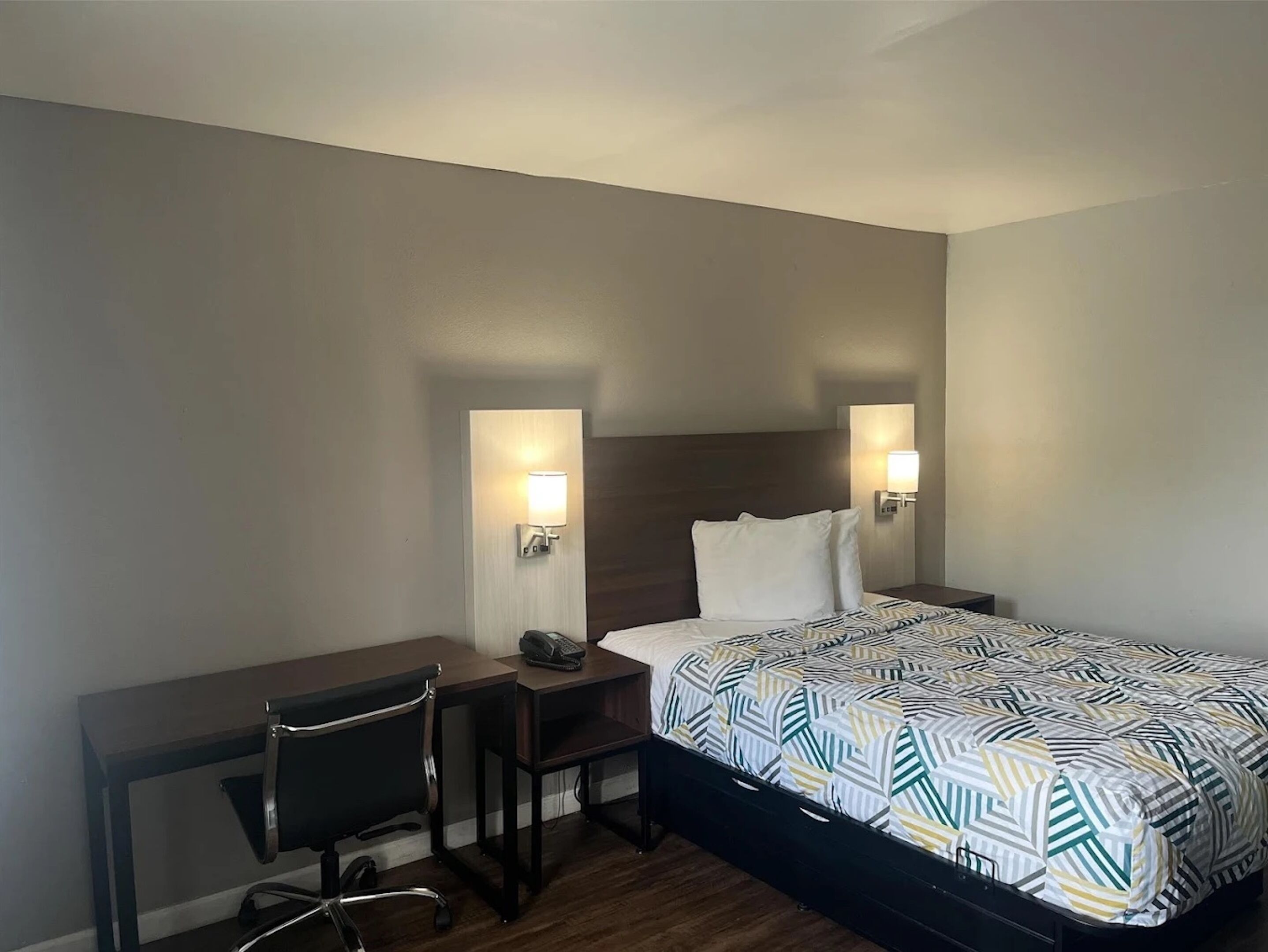 A hotel room with a bed, desk, chair, and two wall-mounted lamps.