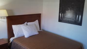 Un lit bien fait, avec des oreillers blancs et une couverture marron à rayures, une table de chevet en bois avec une lampe, et une photographie en noir et blanc d’un chemin bordé d’arbres accrochée au mur.