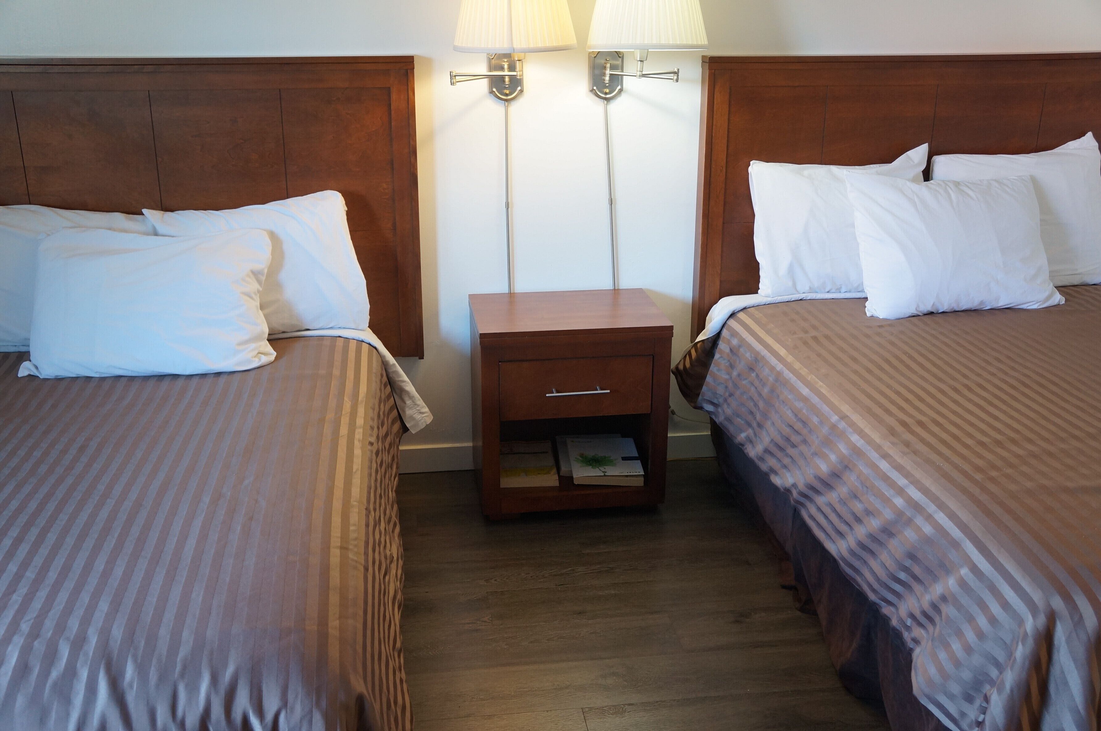 A hotel room with two beds, each with white linens, a wooden nightstand with a drawer, and two wall-mounted lamps.