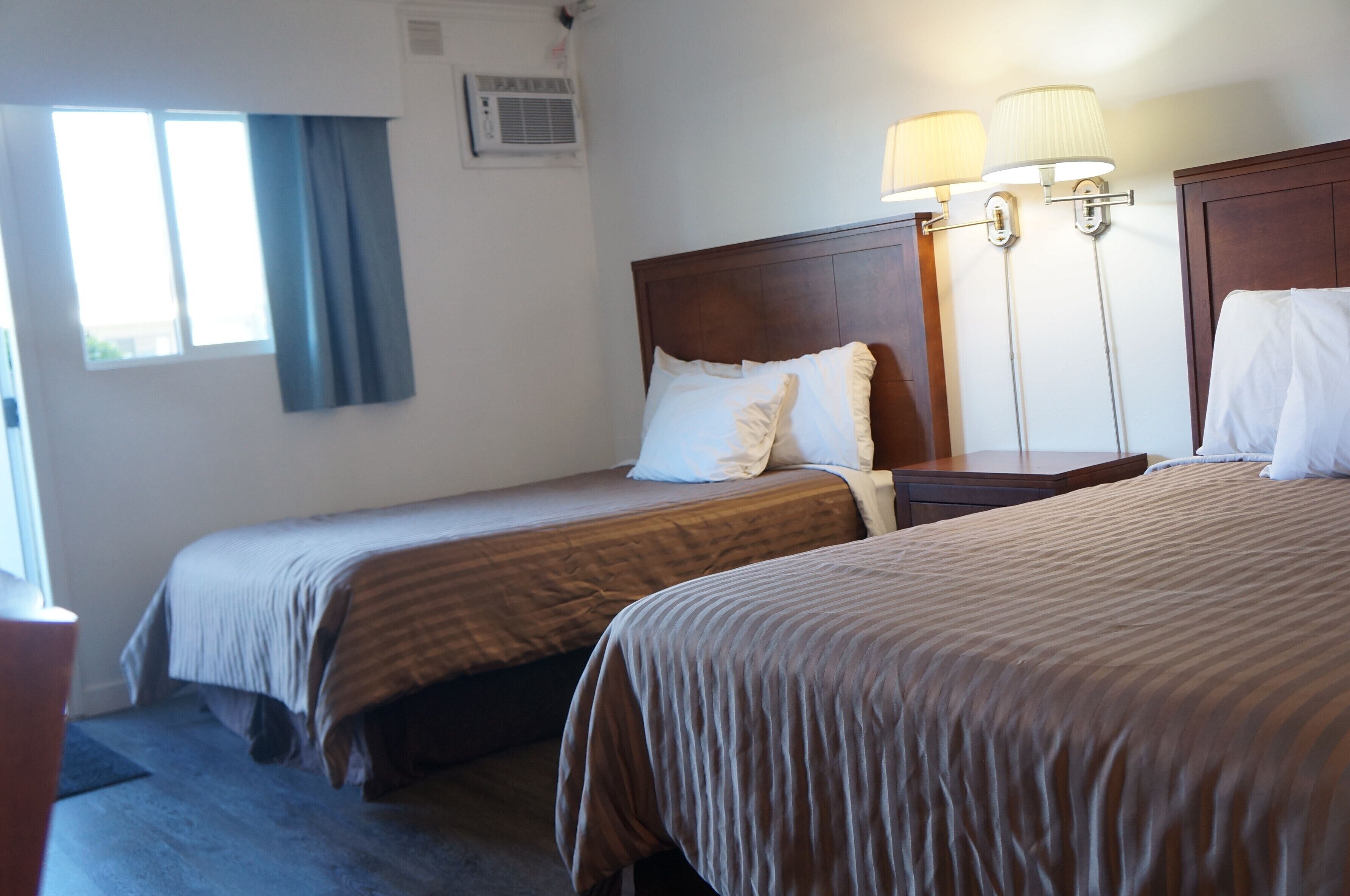A hotel room with two single beds, a wooden headboard, a nightstand, and a lamp.