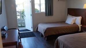 Habitación de hotel con dos camas, un escritorio y una silla. Se ve una ventana con cortinas, una puerta que da al exterior y un pequeño equipo de aire acondicionado en la pared.