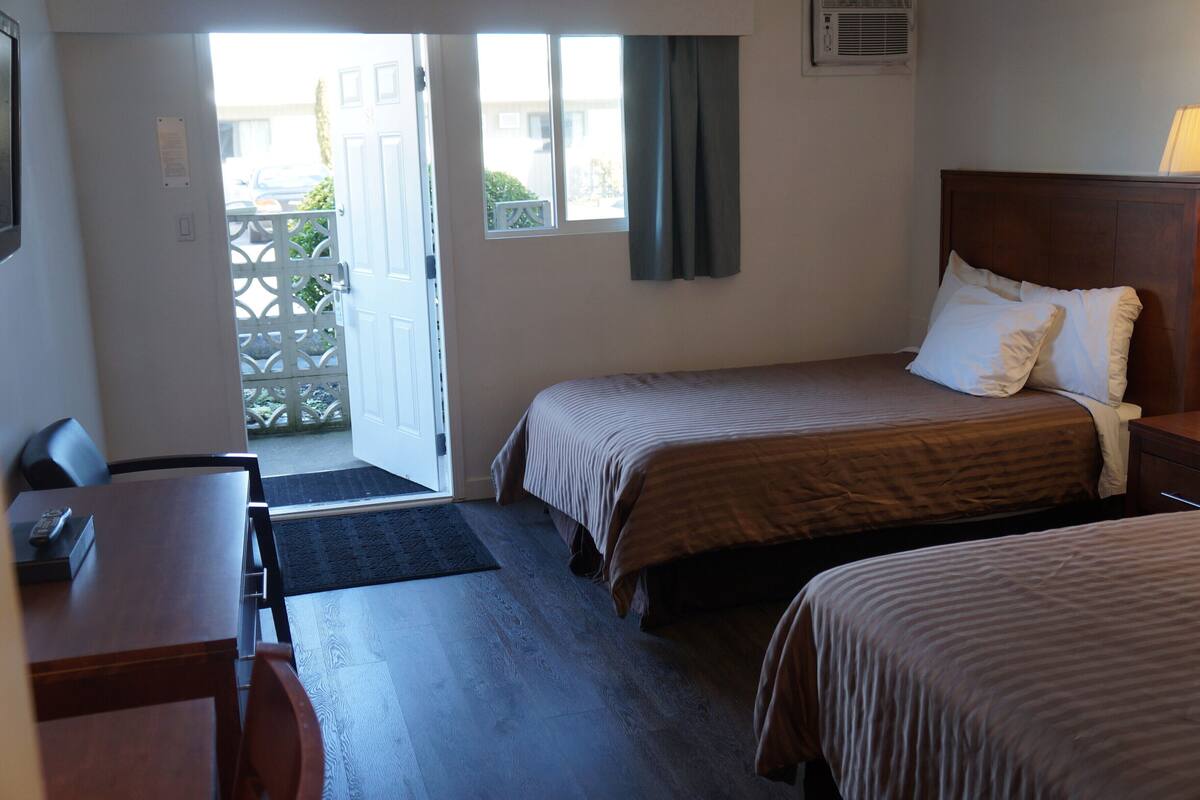 Habitación de hotel con dos camas, un escritorio y una silla. Se ve una ventana con cortinas, una puerta que da al exterior y un pequeño equipo de aire acondicionado en la pared.