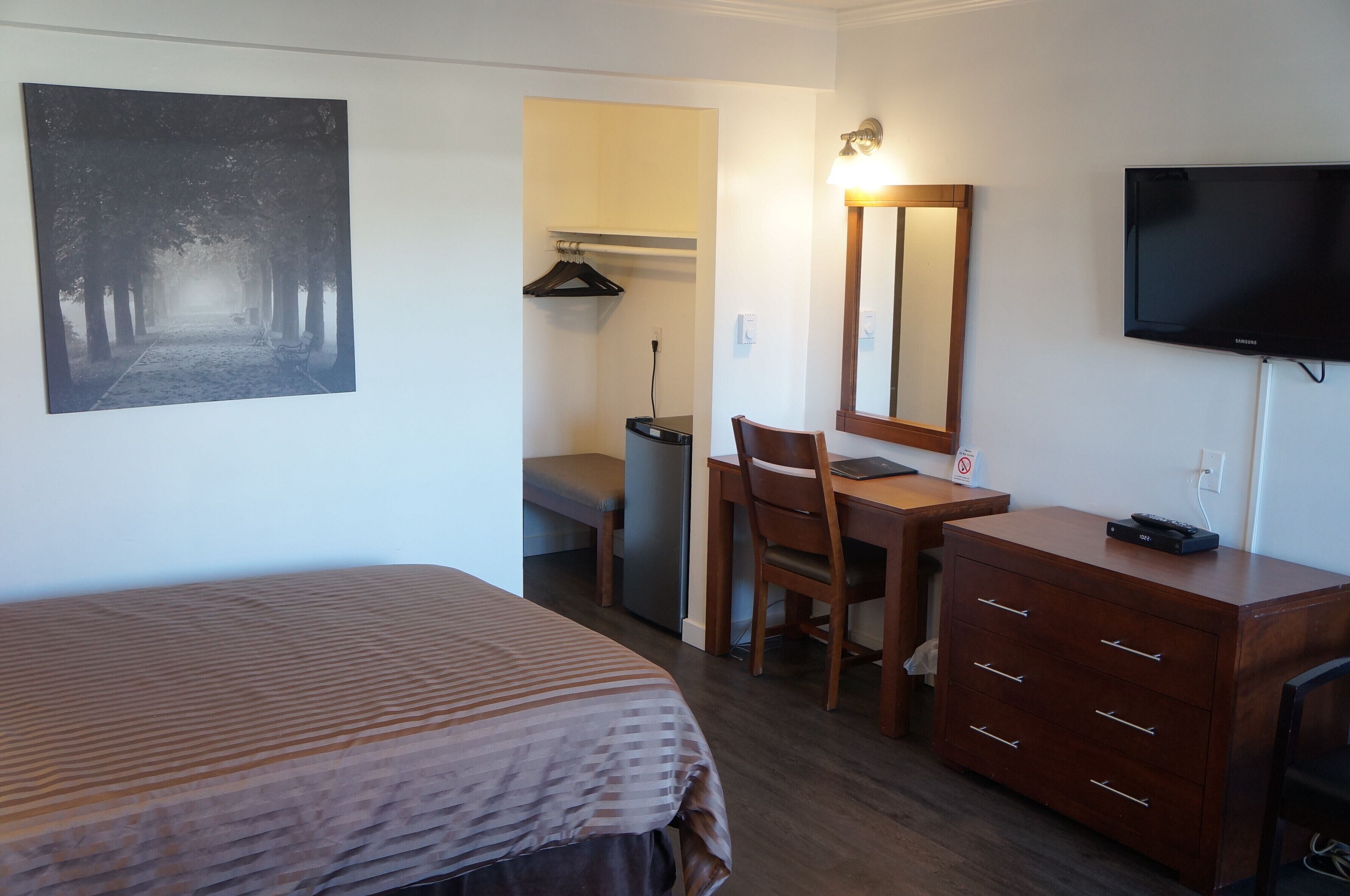 A hotel room with a bed, a desk with a chair, a dresser, a TV, and a painting on the wall.