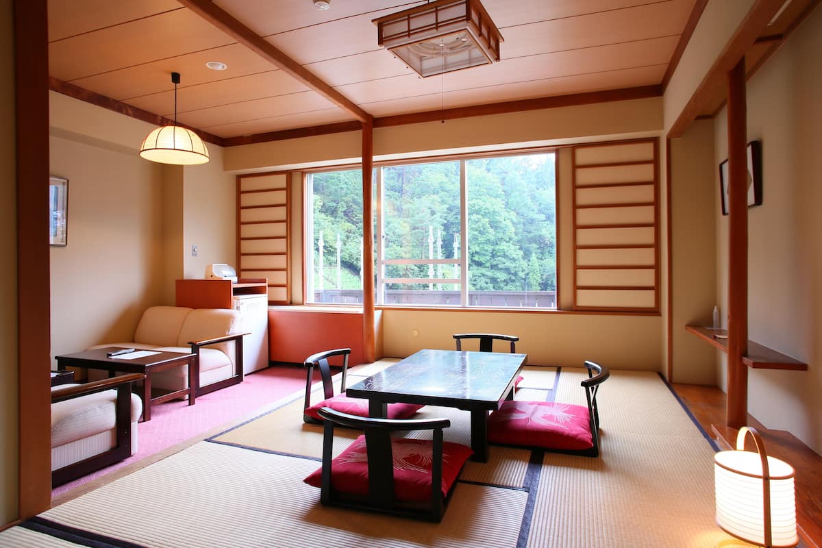 A traditional Japanese room with tatami flooring, a low table with cushions, and a view of a forest through large windows.