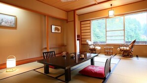 A traditional Japanese room with tatami flooring, a low table with chairs, a sliding door, and a view of greenery outside. - Nozawa Grand Hotel (Nozawaonsen)