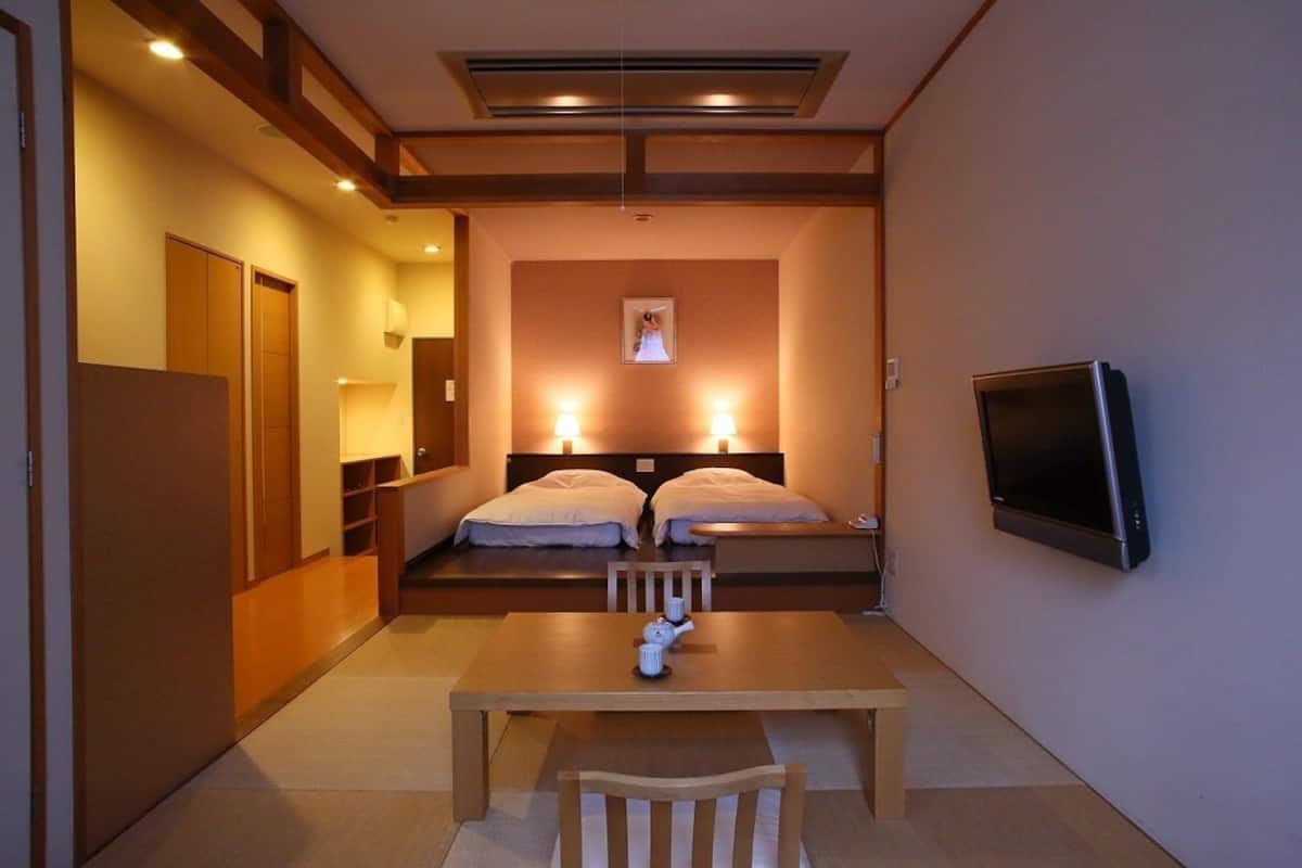 A hotel room with a wooden table, two beds with white linens, a flat-screen TV mounted on the wall, and a framed picture above the beds.