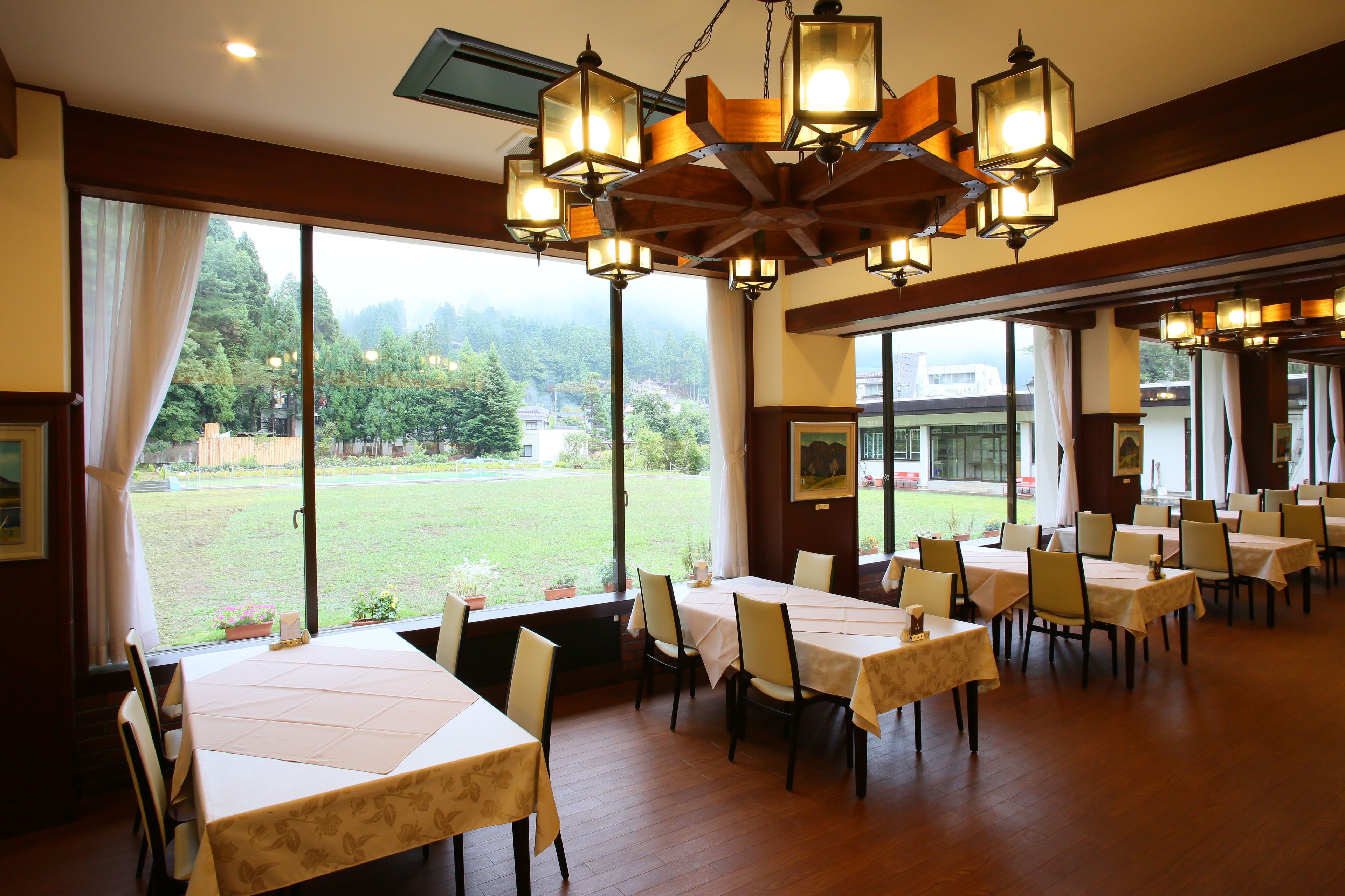 A dining area with multiple tables and chairs, large windows with a view of a green landscape, and a decorative ceiling light fixture.