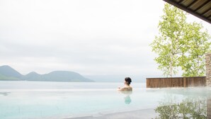Une personne se trouve dans une piscine à débordement aux eaux cristallines, avec une vue sur des montagnes lointaines et une structure en bois.