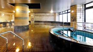 A spacious indoor swimming pool with a tiled floor, a central column, and a seating area with a view of the outside.