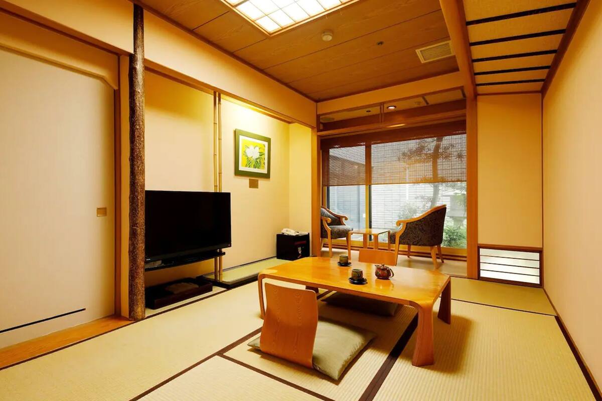 Ein traditioneller japanischer Raum mit Tatami-Böden, einem Fernseher, einem Tisch mit Stühlen und einem großen Fenster.