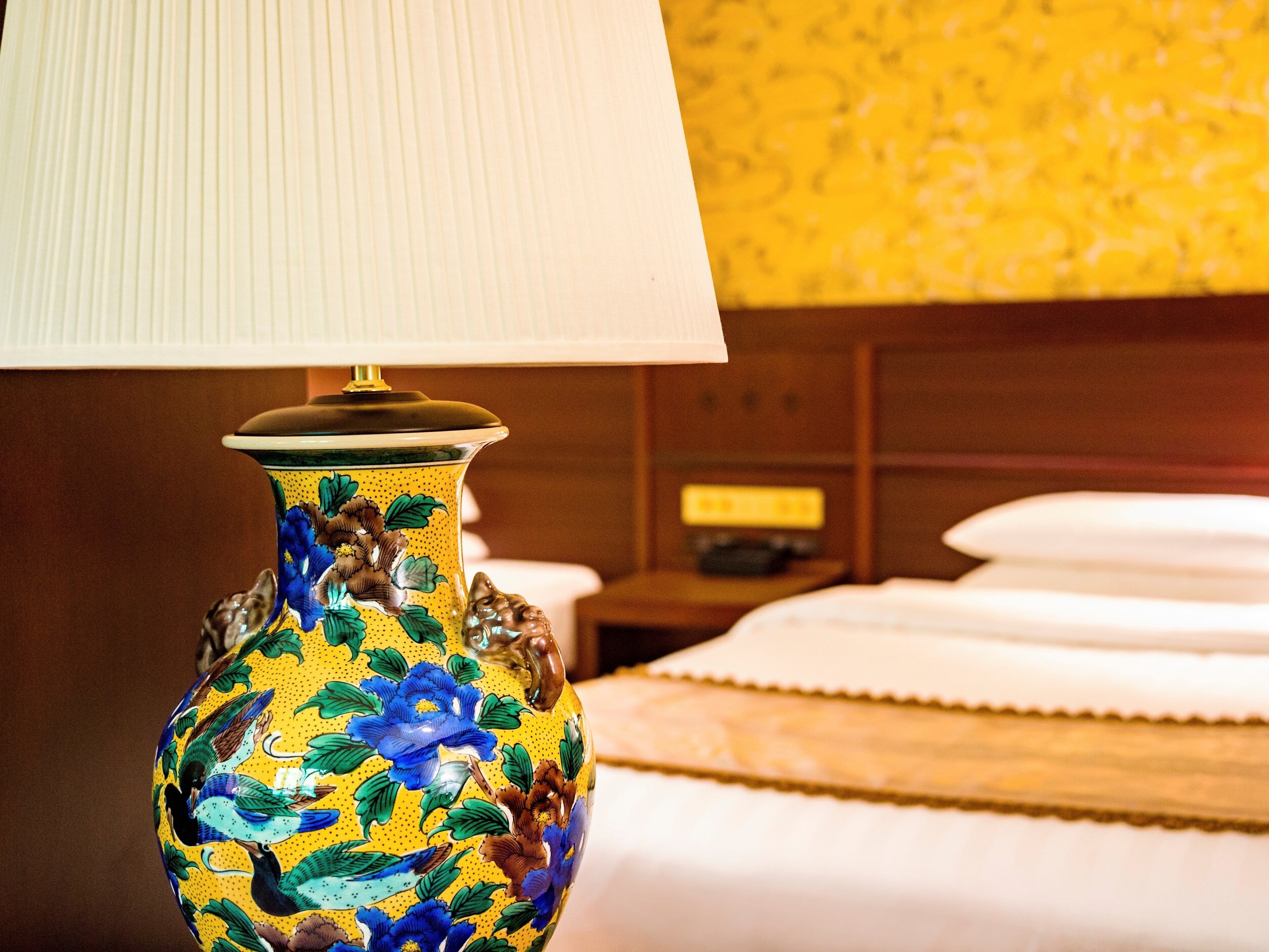 A decorative vase with floral patterns, a bed with white linens, and a nightstand with a lamp.
