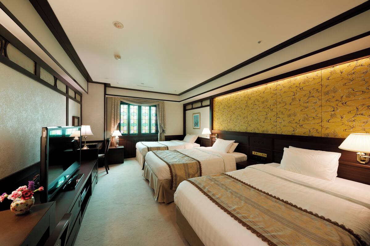 Ein Hotelzimmer mit drei Betten, einem Fernseher, einem Schreibtisch mit Lampe und einem großen Fenster.