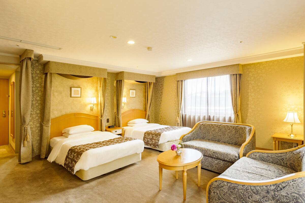 Ein Hotelzimmer mit zwei Betten, einem Schlafsofa, einem kleinen Tisch und einem Fenster mit Vorhängen.