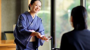 A person in traditional attire serving food to another individual in a room with a large window.
