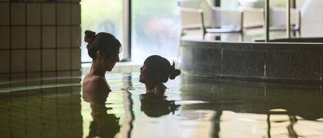 An indoor swimming pool with clear water, a tiled wall, and a view of outdoor seating.
