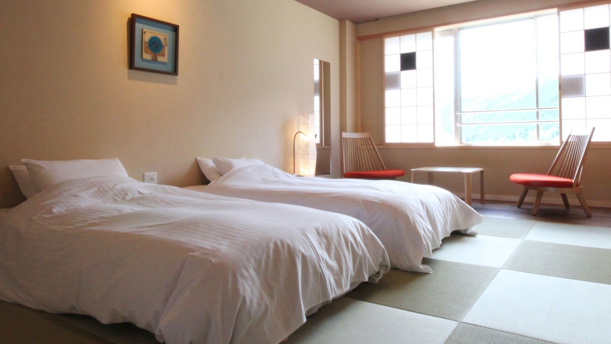Une chambre avec deux lits, une petite table, deux chaises, un tableau au mur et une fenêtre donnant sur un paysage agréable.