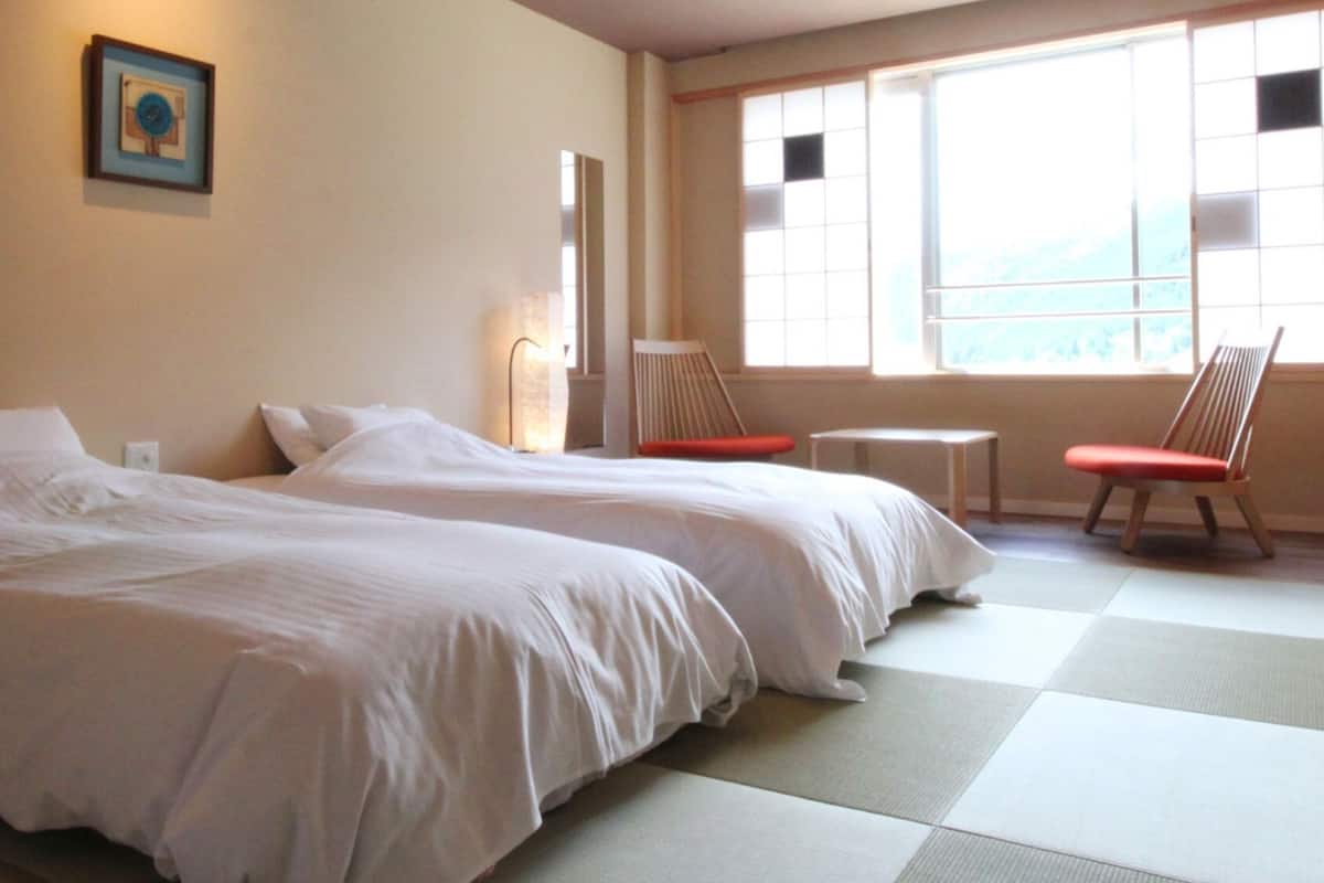 A room with two beds, a small table, two chairs, a picture on the wall, and a window with a view.