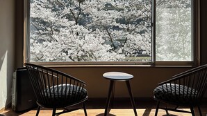Une pièce dotée d’une grande fenêtre donnant sur un cerisier en fleurs. On y trouve deux chaises noires et une petite table ronde.