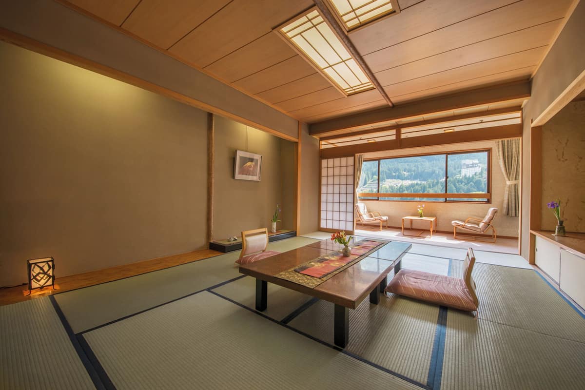 A traditional Japanese room with tatami flooring, a low table, and sliding doors leading to a balcony with a view of a forested area.