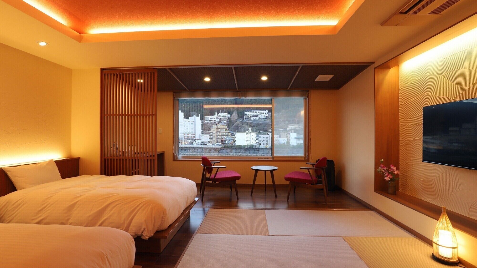 Une chambre d’hôtel comprenant un lit, un coin salon avec des fauteuils, une grande fenêtre donnant sur la ville et un téléviseur à écran plat fixé au mur.
