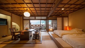 Une pièce de style japonais traditionnel, avec un sol recouvert de tatamis, une table basse entourée de chaises, un lit avec une literie blanche et une vue sur une montagne, visible à travers de grandes portes coulissantes.