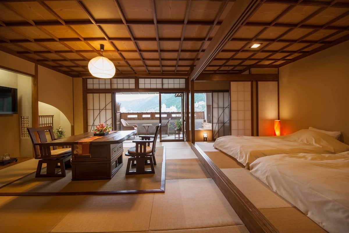 A traditional Japanese-style room with tatami flooring, a low table with chairs, a bed with white bedding, and a view of a mountain through large sliding doors.