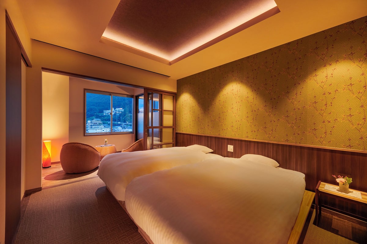 A hotel room with two beds, a large window with a city view, a wall-mounted lamp, and a small table with a plant.