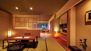 Um quarto tradicional japonês com tatames, uma mesa baixa com almofadas, TV na parede e uma porta deslizante com vista para a cidade.