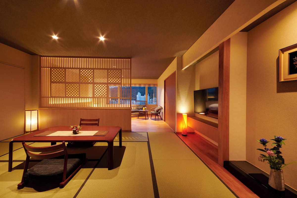 A traditional Japanese-style room with tatami flooring, a low table with cushions, a TV mounted on the wall, and a sliding door with a view of the cityscape.