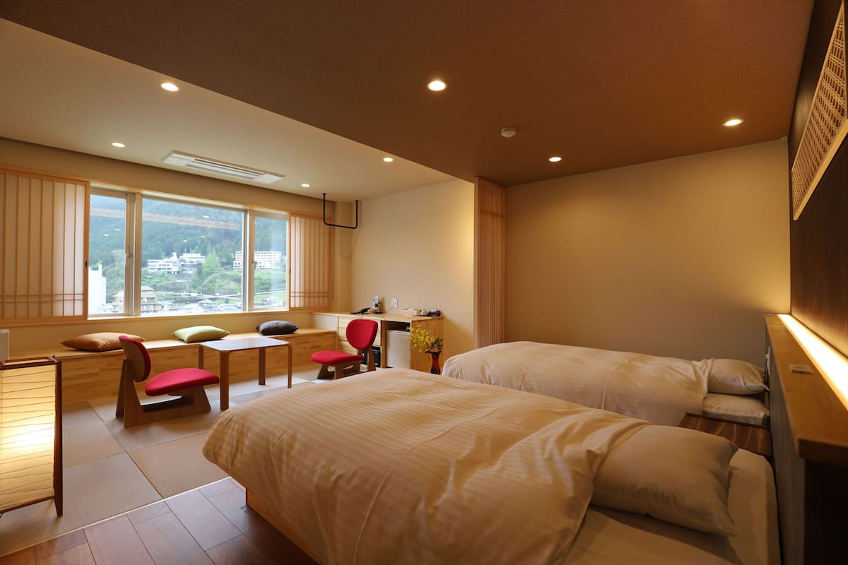A hotel room with two beds, a desk, a chair, and a view of a cityscape through large windows.