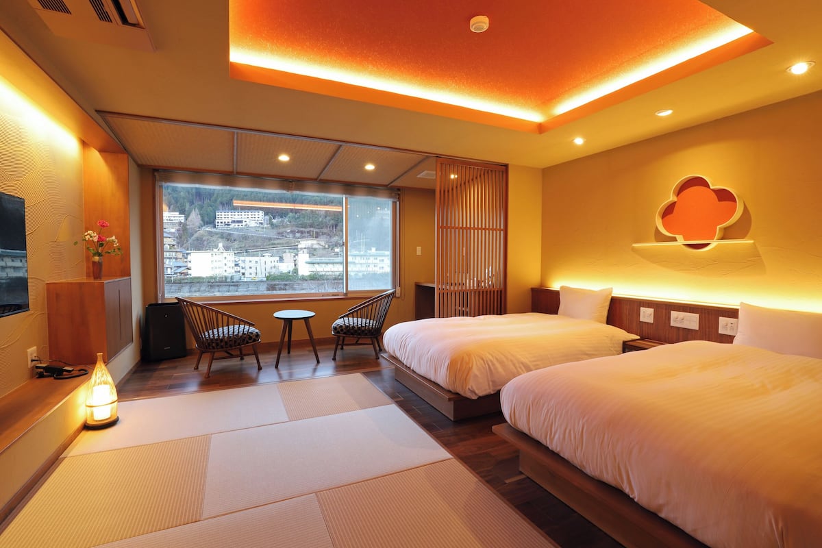 A hotel room with two beds, a sitting area with a chair and table, a view of a cityscape, and a decorative wall piece.