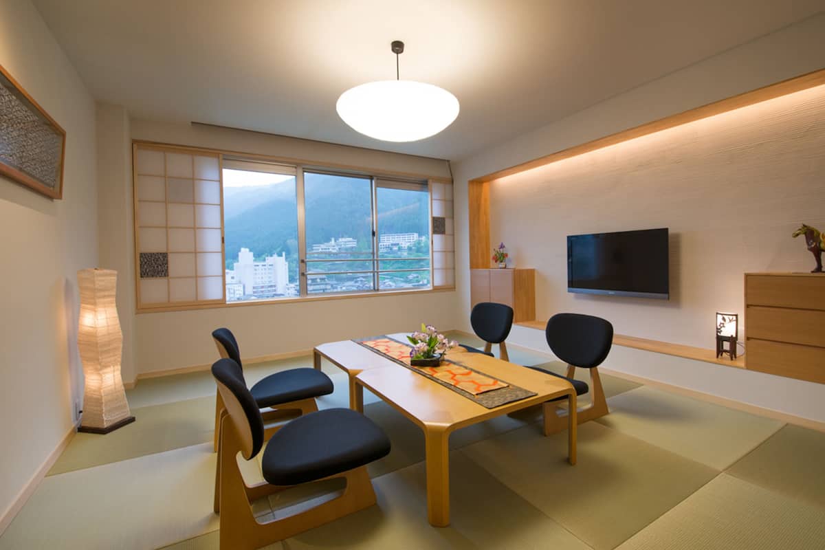 Ein traditioneller japanischer Raum mit Tatami-Boden, einem niedrigen Tisch mit Kissen, einem Fernseher und Blick auf eine Stadtlandschaft durch große Fenster.