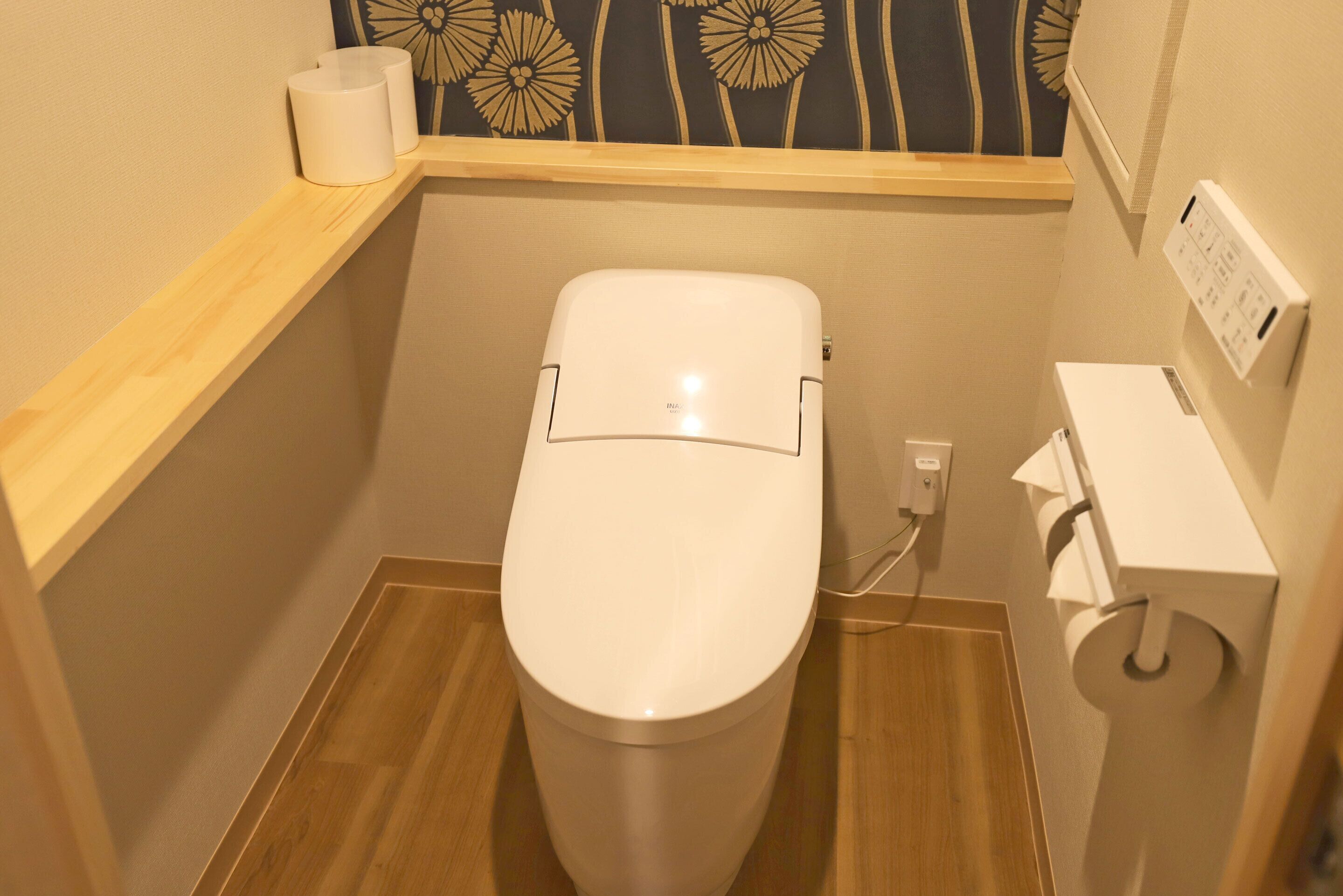 A small, compact bathroom with a white toilet, a wooden shelf with a roll of toilet paper, and a wall-mounted electrical outlet.