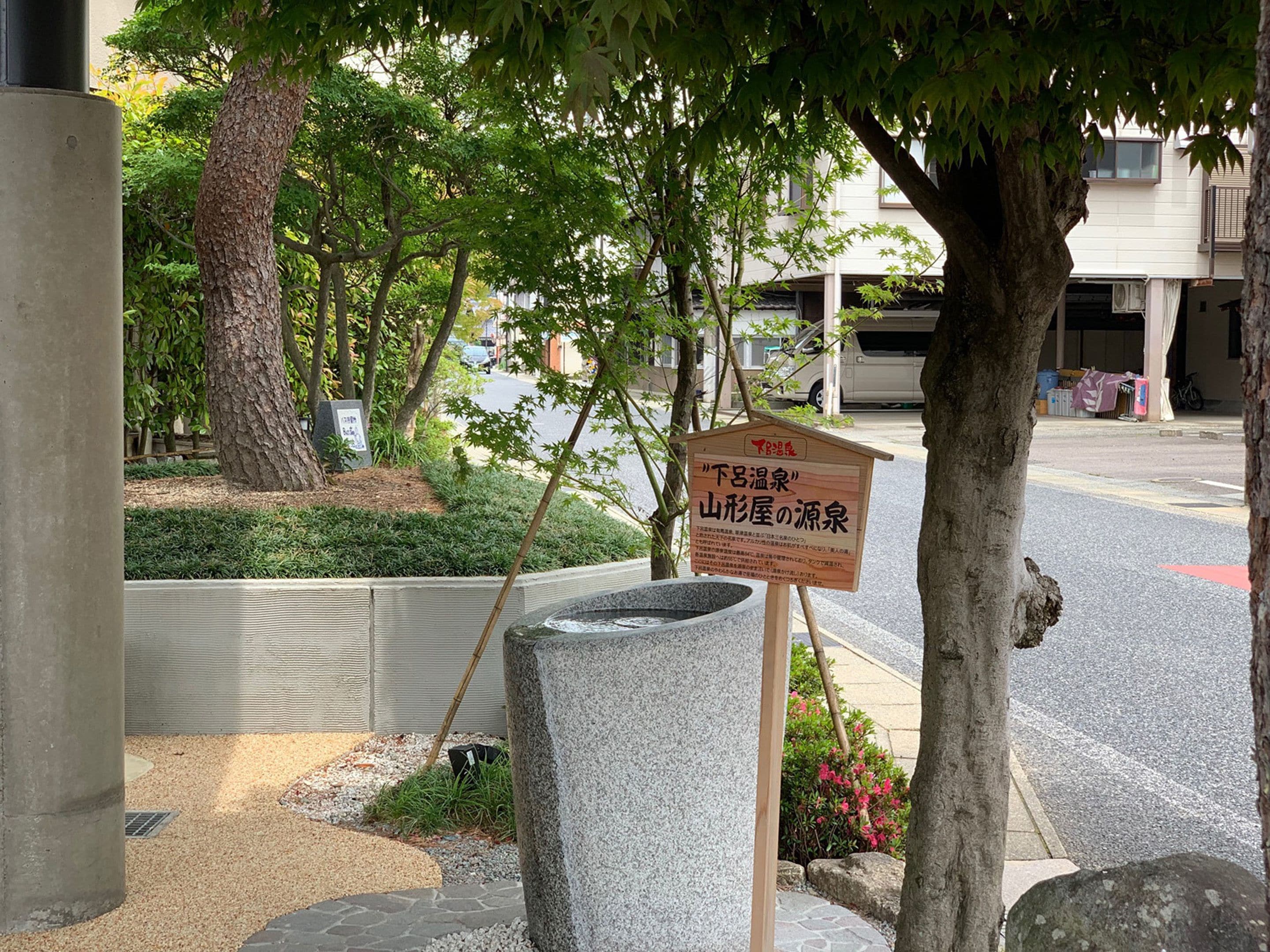 A stone water fountain with a sign in Japanese, surrounded by trees and a sidewalk.