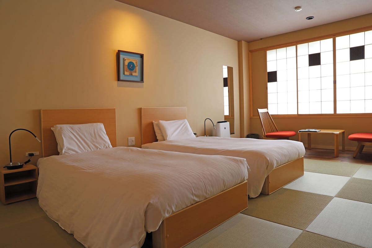 A hotel room with two beds, a bedside table with a lamp, a chair, a small table, and a large window with a view.