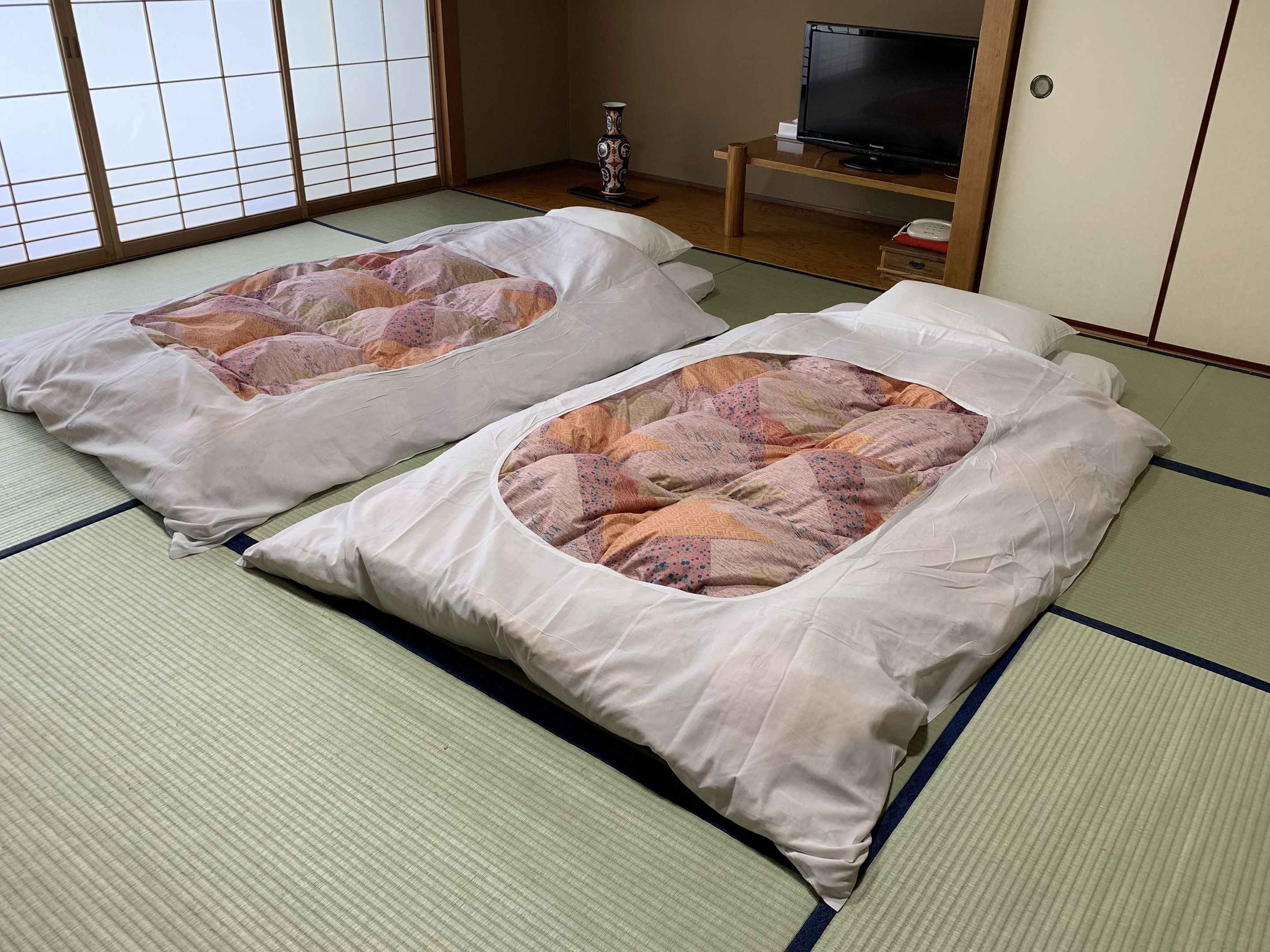 Two tatami mat beds with pillows, a television, and a traditional Japanese vase in a room with sliding doors.