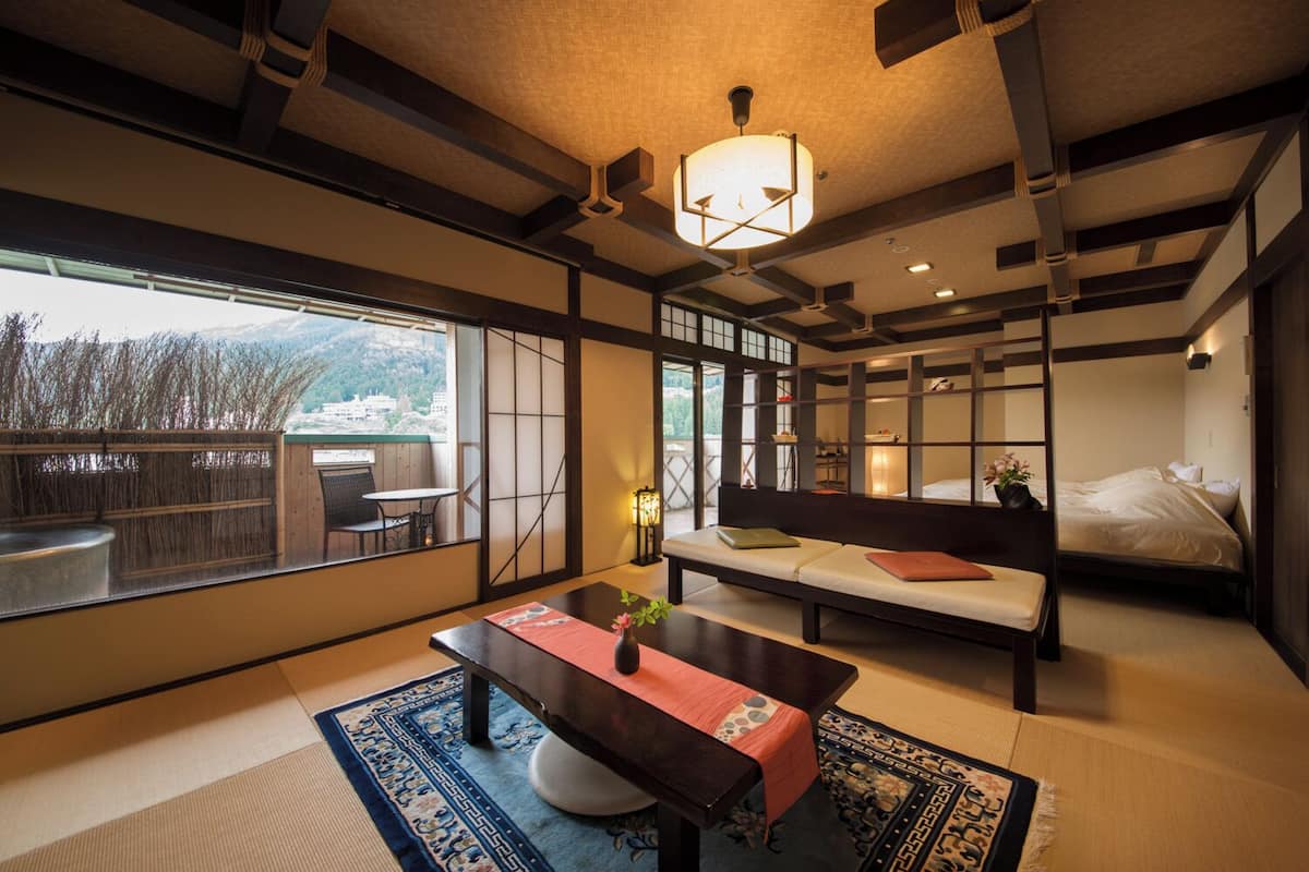 A traditional Japanese-style room with tatami flooring, a low table with a red table runner, a sofa with cushions, and a large window offering a view of the outdoors.