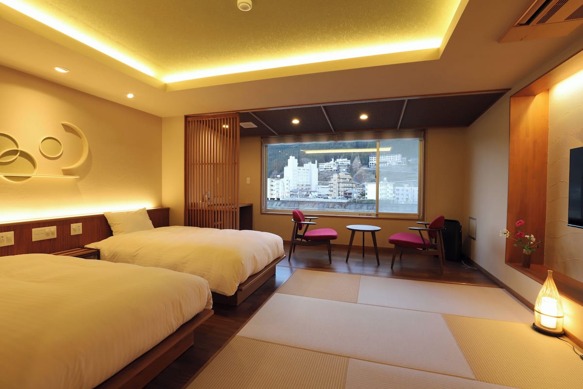 A hotel room with two beds, a city view, a small table, and a chair.