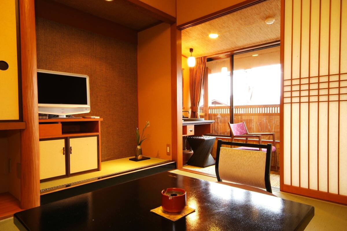 Une pièce de style japonais traditionnel, avec un sol recouvert de tatamis, une petite table sur laquelle se trouve une tasse rouge, un téléviseur posé sur un meuble en bois, et des portes coulissantes donnant sur un balcon d’où l’on peut admirer une clôture en bois.