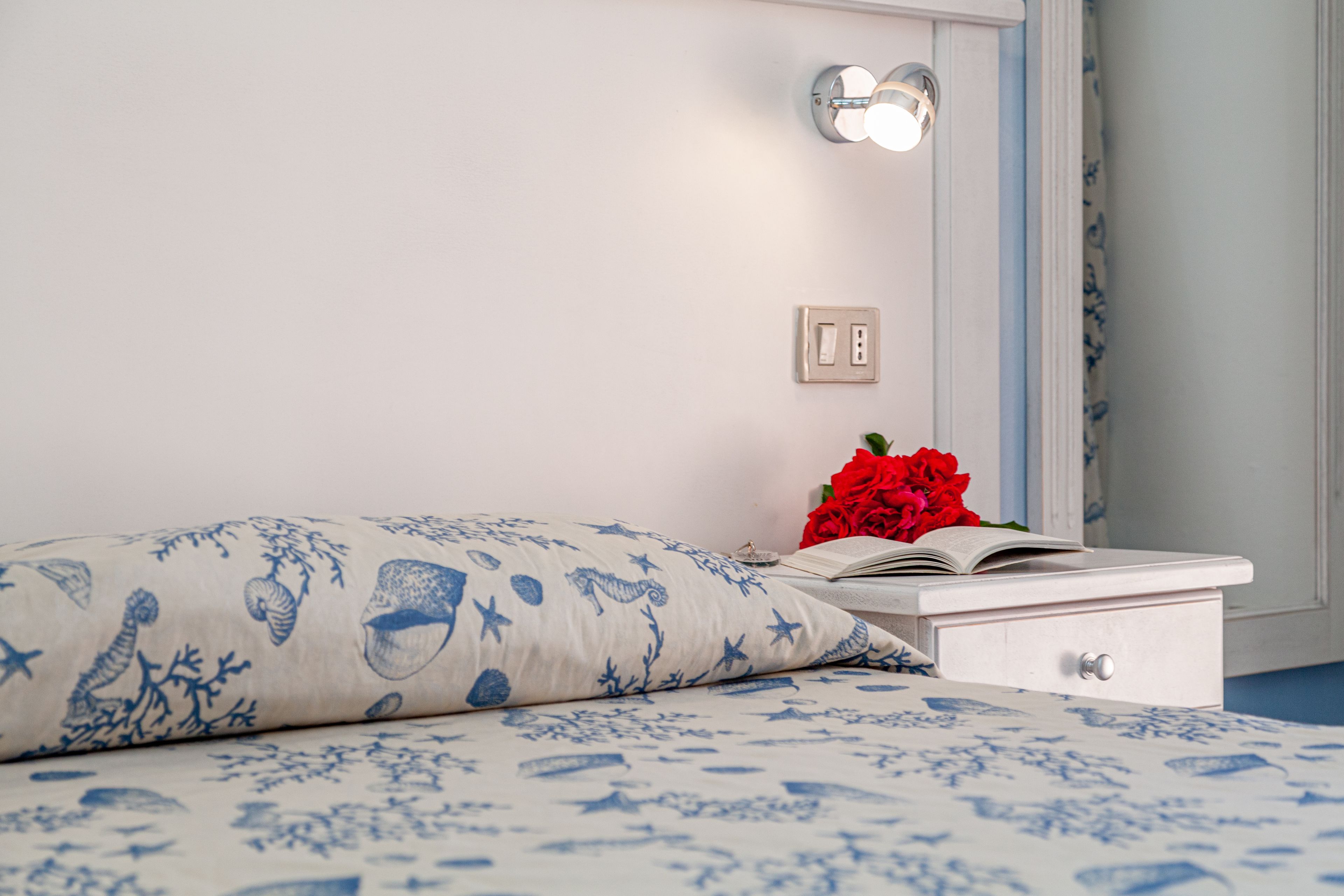 Un letto rifatto con cura, con copriletto blu e bianco a fantasia, un comodino bianco con un libro e un fiore rosso, e un lampadario fissato al muro.